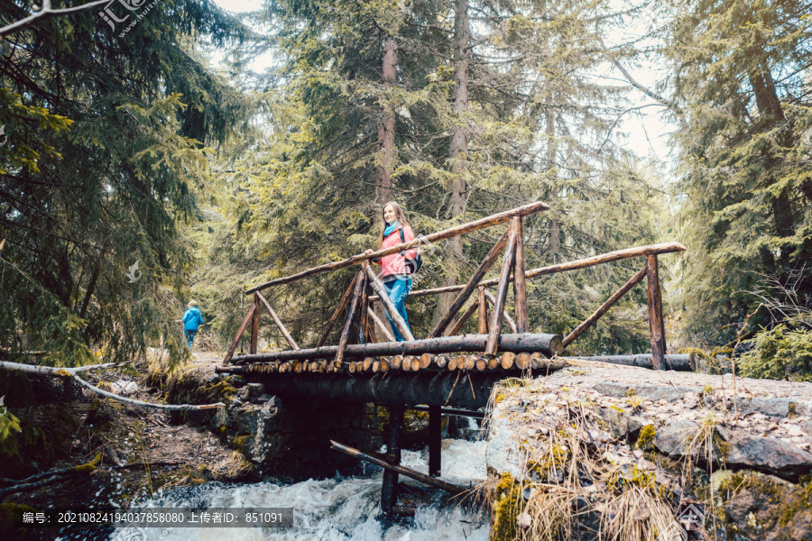 一个徒步旅行的女人站在树林里的桥上，看着山间小溪的水
