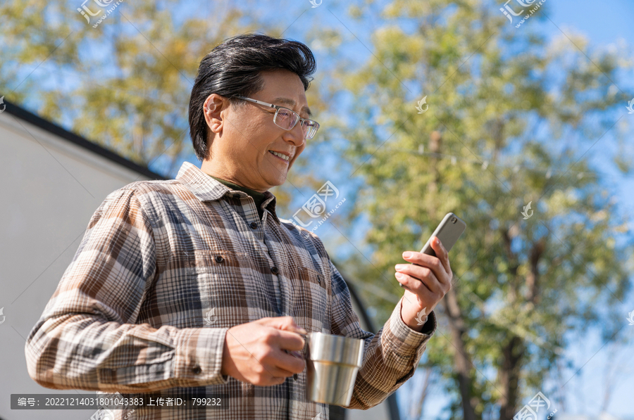 一个老年男人站在房车前看着手机视频喝咖啡